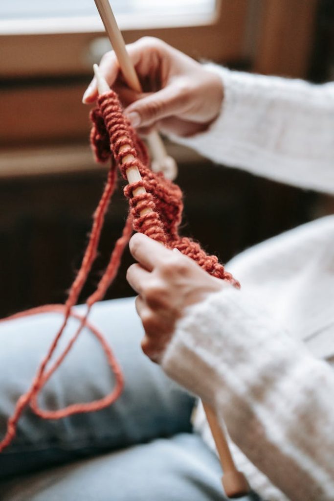 Close-up of a woman knitting a scarf with red wool indoors, emphasizing relaxation and craftsmanship.